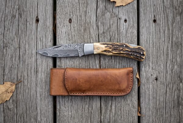 Damascus folding pocket knife with stag horn handle lying open on grey boards from above, leather sheath below it