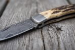 Damascus folding pocket knife in close view at the steel bolster, stag horn handle and patterned blade on old wood