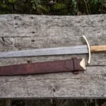 Damascus longsword with olive wood grip resting above its brown leather scabbard with brass tips on a weathered table