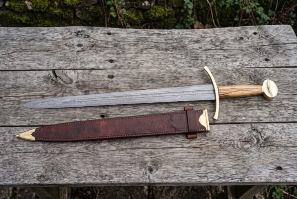 Damascus longsword with olive wood grip resting above its brown leather scabbard with brass tips on a weathered table