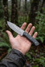 Damascus skinner knife with black micarta handle resting on an open palm in a forest, the blade pattern clearly visible