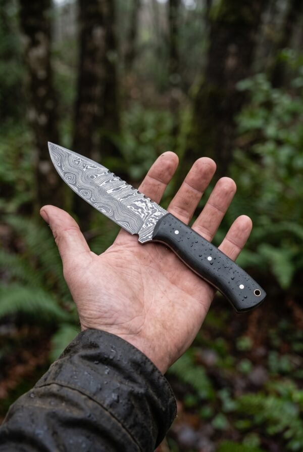 Damascus skinner knife with black micarta handle resting on an open palm in a forest, the blade pattern clearly visible