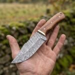 Damascus skinner knife with wood handle held flat in an open palm outdoors, blade pattern facing the camera