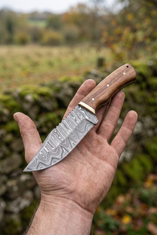 Damascus skinner knife with wood handle held flat in an open palm outdoors, blade pattern facing the camera