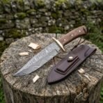 Damascus steel bowie knife with wood handle and brass guard on a tree stump with wood chips, brown leather sheath beside it