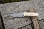 Damascus steel bull cutter knife with pale camel bone handle laid across a strip of bark on an old wooden table