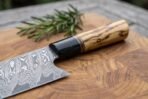 Damascus steel chef knife with olive wood handle and black horn ferrule, close up of the heel on an end grain board with rosemary