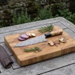Damascus steel chef knife with olive wood handle on an end grain board with sliced red onion, rosemary and a brown leather sheath