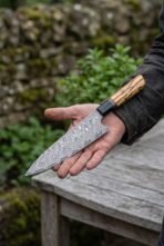 Damascus steel chef knife with olive wood handle resting across an open palm in a garden beside a weathered wooden table