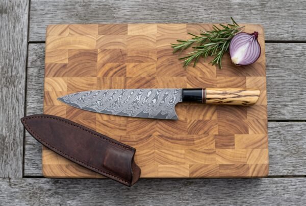 Damascus steel chef knife with olive wood handle seen from above on an end grain board next to its brown leather sheath and a halved onion