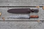Damascus steel dagger with black guard and reddish wood handle laid flat on grey boards below its brown leather sheath