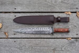 Damascus steel dagger with black guard and reddish wood handle laid flat on grey boards below its brown leather sheath