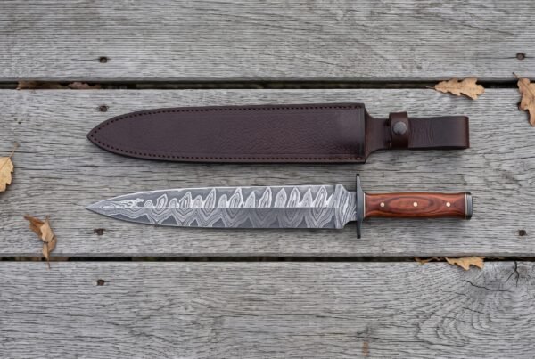 Damascus steel dagger with black guard and reddish wood handle laid flat on grey boards below its brown leather sheath