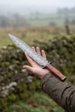 Damascus steel dagger with wood handle and brass pins resting on an open palm in front of a misty green field and stone wall