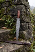 Damascus steel double edged dagger with reddish wood handle standing against a mossy stone wall above its brown leather sheath