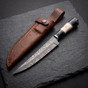 Damascus steel fillet knife with bone and black horn handle beside its brown leather sheath on a dark slate board