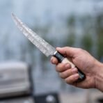 Damascus steel fillet knife with bone and black horn handle held in hand outdoors with a blurred lakeside behind