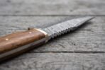Close view of a Damascus steel knife blade with serrated spine and wood handle with brass guard on grey boards