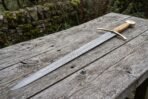 Damascus steel longsword with olive wood grip and brass fittings laid full length across a rustic table by a stone wall