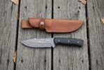 Damascus steel skinner knife with black micarta handle laid flat on wooden boards below its tan leather sheath