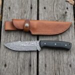 Damascus steel skinner knife with black micarta handle laid flat on wooden boards below its tan leather sheath