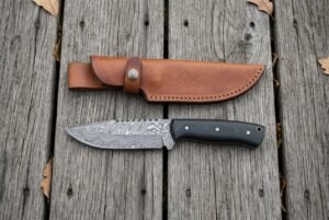 Damascus steel skinner knife with black micarta handle laid flat on wooden boards below its tan leather sheath