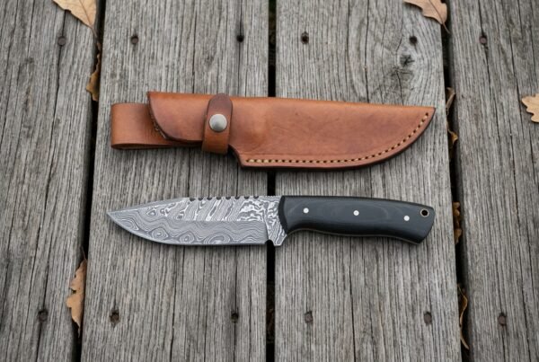 Damascus steel skinner knife with black micarta handle laid flat on wooden boards below its tan leather sheath
