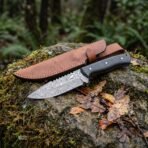 Damascus steel skinner knife with black micarta handle on a mossy rock beside its tan leather sheath and fallen leaves