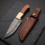 Damascus steel skinner knife with camel bone and wood handle beside its brown leather sheath on dark slate