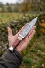 Double edged Damascus dagger with wood and bone handle held on an open palm beside a mossy stone wall