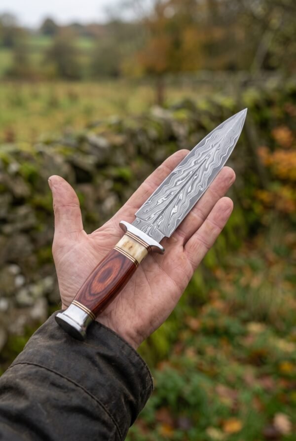 Double edged Damascus dagger with wood and bone handle held on an open palm beside a mossy stone wall