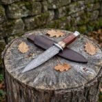Double edged Damascus dagger with wood and bone handle lying across its dark leather sheath on a tree stump