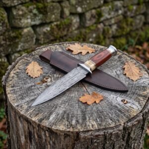 Double edged Damascus dagger with wood and bone handle lying across its dark leather sheath on a tree stump