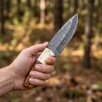 Hand holding a Damascus skinner knife with camel bone and wood handle against a blurred forest background