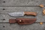 Overhead flat lay of a Damascus skinner knife with wood handle above its brown leather sheath on grey planks