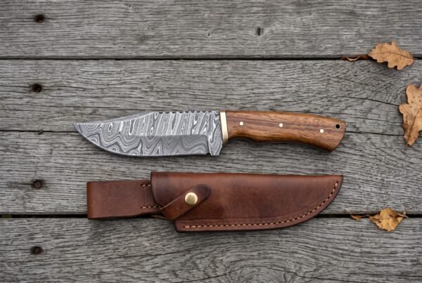 Overhead flat lay of a Damascus skinner knife with wood handle above its brown leather sheath on grey planks