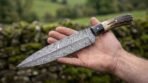 Open hand holding a Damascus dagger with stag horn grip and black horn spacer, green fields blurred behind