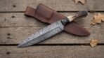 Damascus dagger with stag horn handle crossed over its stitched brown leather sheath on weathered planks with oak leaves