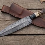 Damascus dagger with stag horn handle crossed over its stitched brown leather sheath on weathered planks with oak leaves