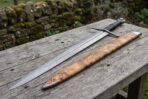 Wide view of a Damascus dagger sword and olive burl wood scabbard lying side by side across a long wooden table