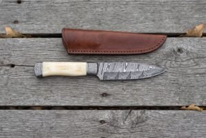 Damascus knife with camel bone handle lying below its brown leather sheath on grey weathered planks from above