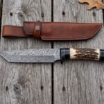 Damascus tanto knife with stag horn and black horn handle laid beside its brown leather sheath on weathered wooden boards
