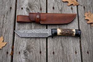 Damascus tanto knife with stag horn and black horn handle laid beside its brown leather sheath on weathered wooden boards
