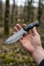 Damascus tracker knife with black horn handle and serrated spine held in an open hand in the forest