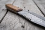Close view of a Damascus tracker knife with saw back spine and hardwood handle lying on grey wooden boards