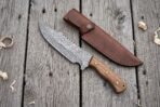 Overhead view of a Damascus tracker knife with hardwood handle beside its brown leather sheath and wood curls