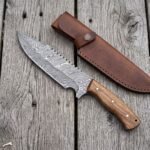 Overhead view of a Damascus tracker knife with hardwood handle beside its brown leather sheath and wood curls