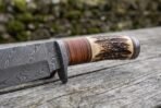 Damascus camp knife close up of the stag horn and stacked leather handle with the dark guard and patterned blade