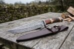 Damascus steel camp knife with stag horn and stacked leather handle lying on a plank table across its brown leather sheath