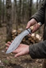 Damascus steel kukri knife with stag horn handle held in both hands in a forest, curved patterned blade in view